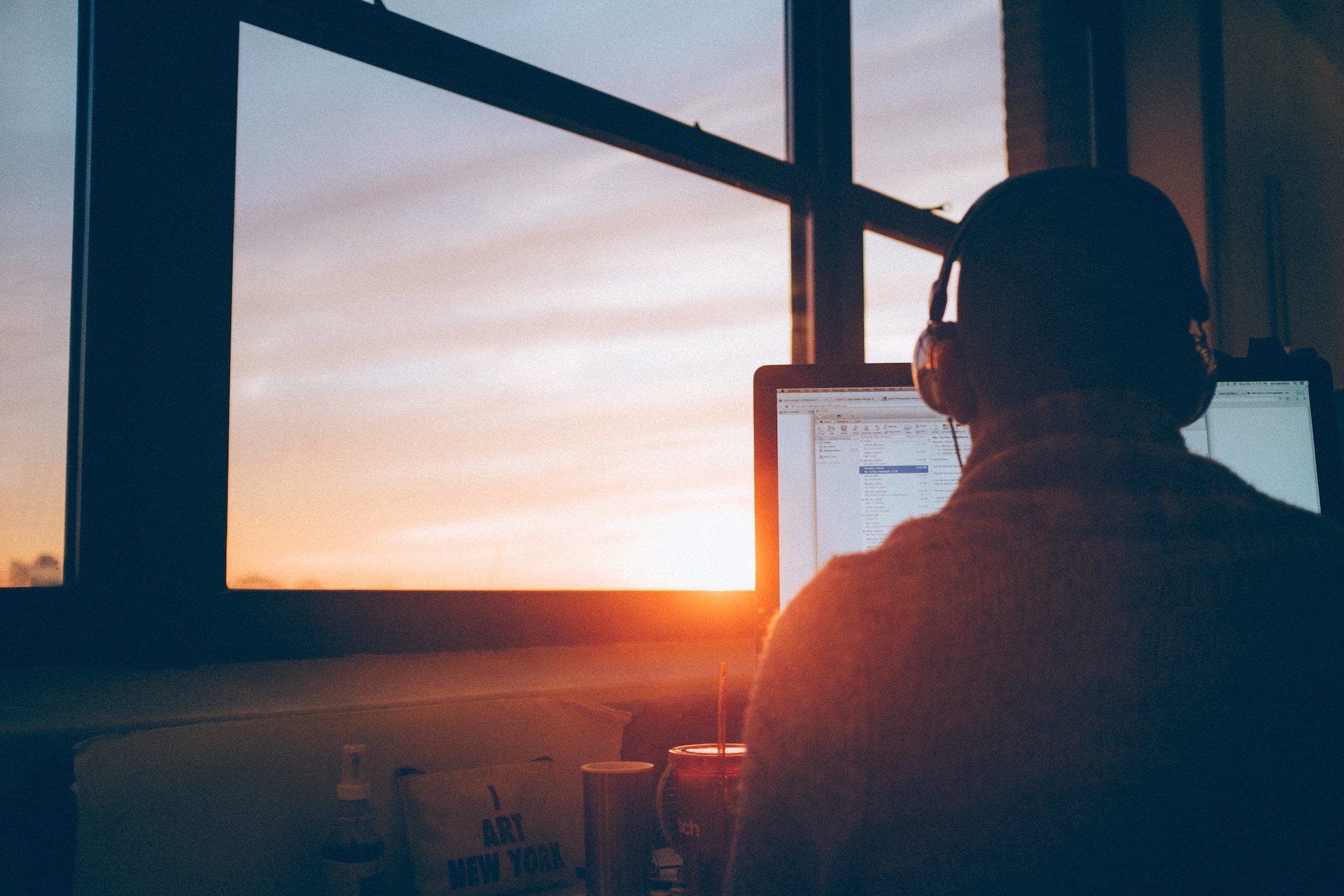 back of a person wearing headphones on a computer next to a window with the sunset shining through
